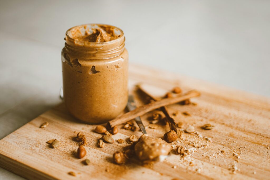 A jar of peanut butter with scattered peanuts and cinnamon on a cutting board.